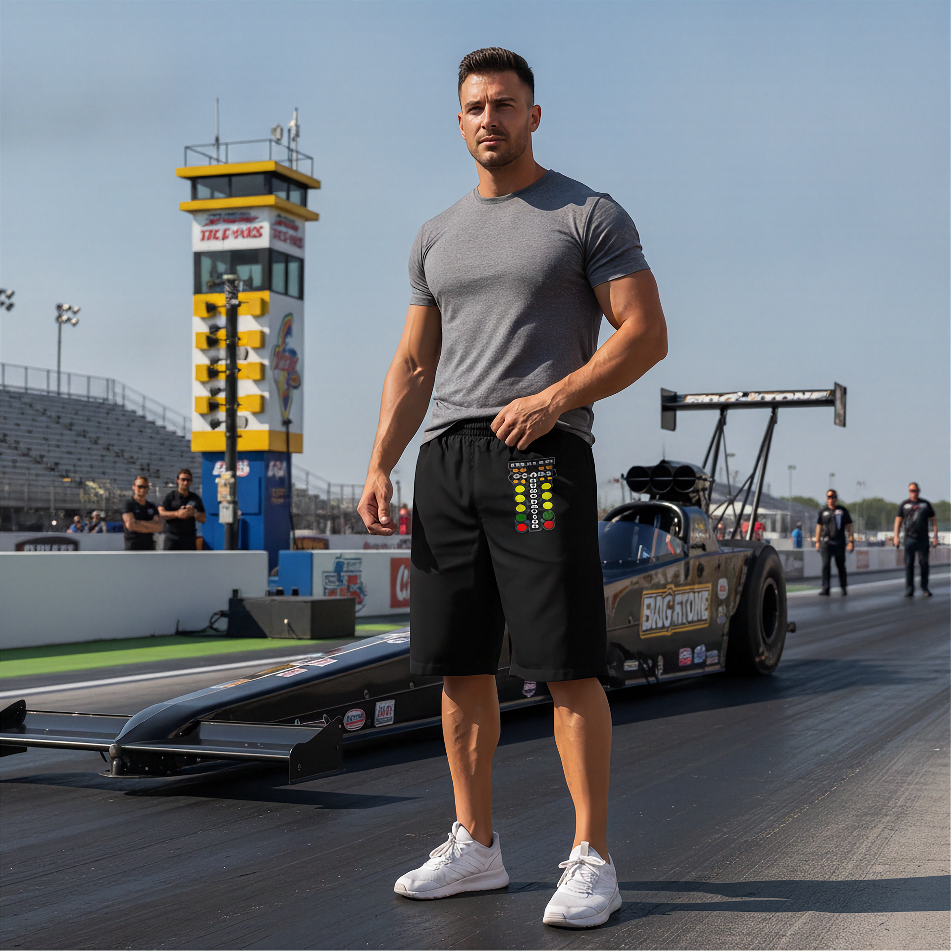 A muscular man stands on a drag racing track wearing a gray t-shirt, black shorts with a colorful graphic on the side, and white sneakers, with a drag race car and a tower in the background.