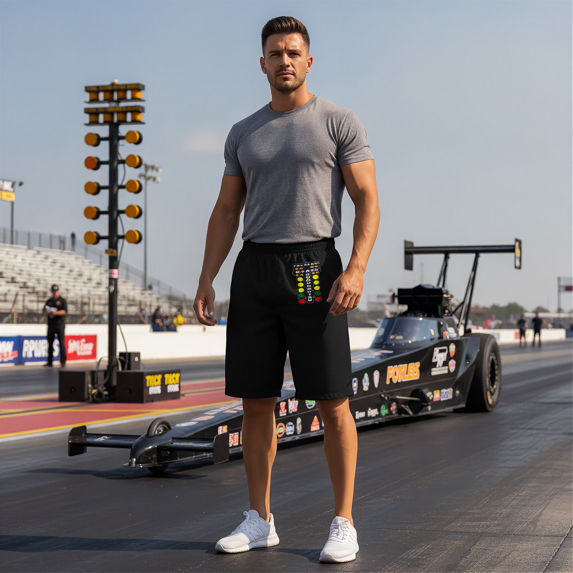 A muscular man stands on a drag racing track wearing a gray t-shirt, black shorts, and white sneakers, with a drag race car and track lights in the background.