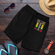 Black athletic shorts with a colorful traffic light graphic on the left leg, styled in a flat lay on a dark wood background with sunglasses, a straw hat, flip-flops, and a watch.