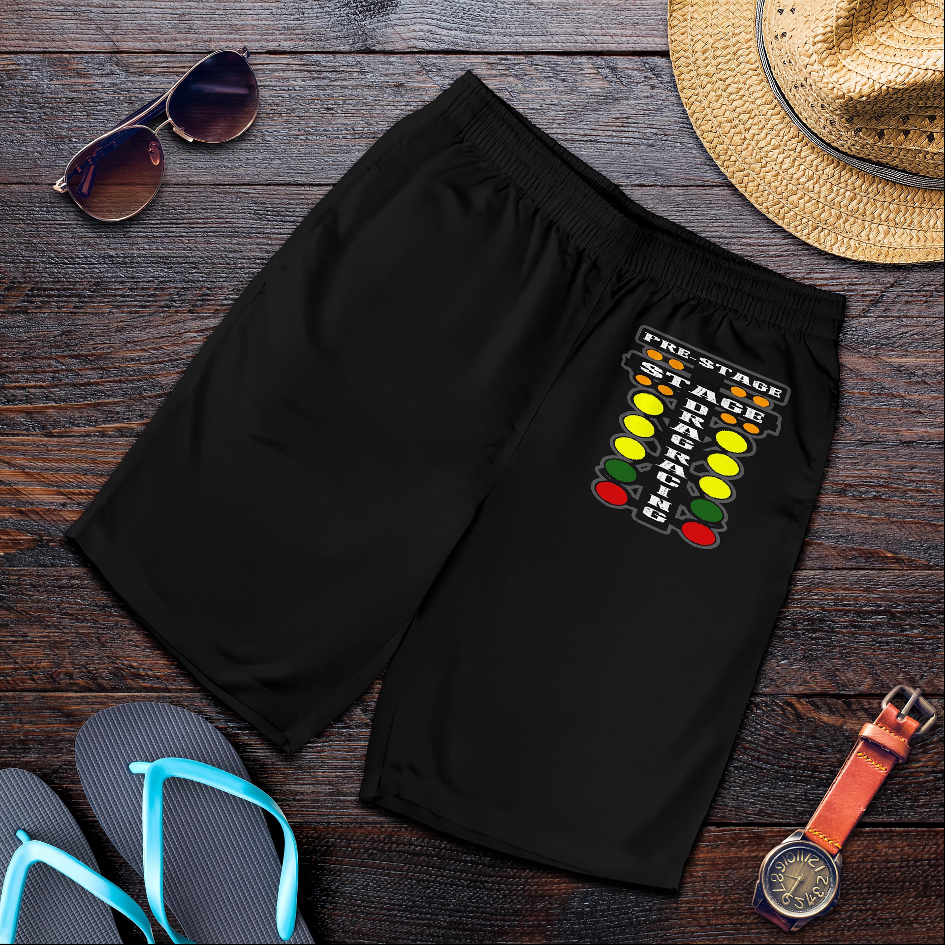 Black athletic shorts with a colorful traffic light graphic on the left leg, styled in a flat lay on a dark wood background with sunglasses, a straw hat, flip-flops, and a watch.