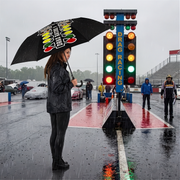 A woman in a black rain jacket and pants stands under a black umbrella with a drag racing logo, holding the handle near a drag racing start line at a wet racetrack with race officials and cars in the background.