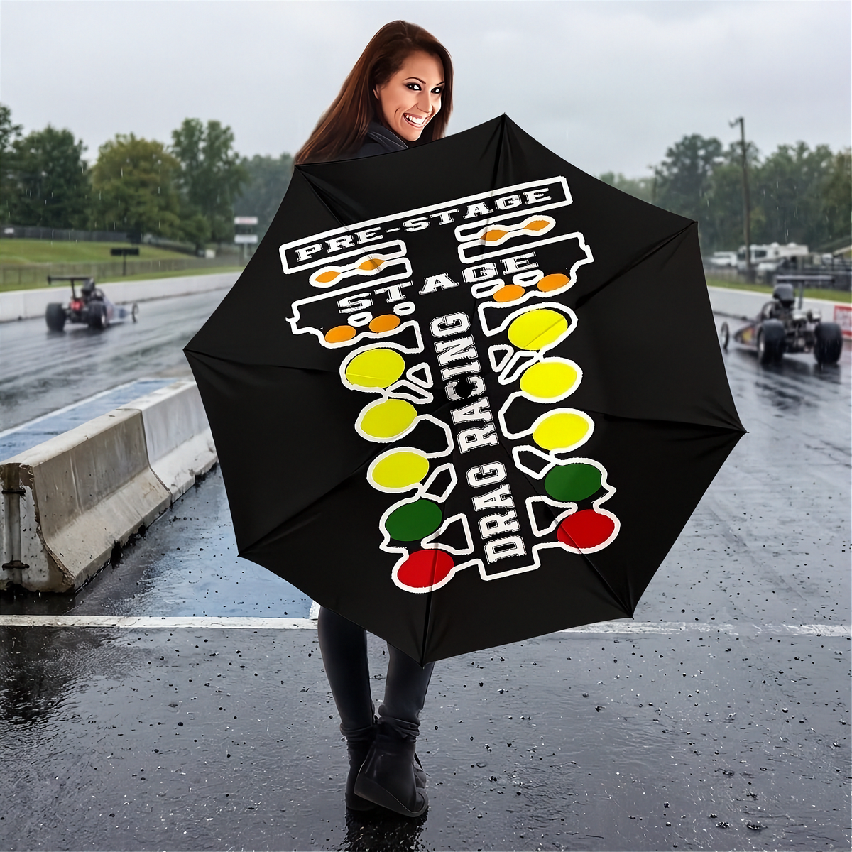 A woman stands on a wet racetrack holding a black umbrella printed with a white, yellow, green, and red drag racing graphic and the words 'PRE-STAGE', 'STAGE', and 'DRAG RACING'.