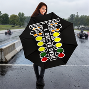 A woman stands on a wet racetrack holding a black umbrella printed with a white, yellow, green, and red drag racing graphic and the words 'PRE-STAGE', 'STAGE', and 'DRAG RACING'.