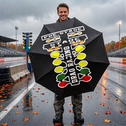 A man standing on a wet racetrack in the rain, holding a large black umbrella printed with white, yellow, green, and red racing graphics and the words 'PRE-STAGE STAGE DRAG RACING'.