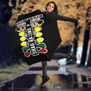 A woman jumps in a puddle outdoors holding a black umbrella with a large white 'Drag Racing' graphic and yellow, green, and red oval accents. The background shows a blurred autumn street scene with trees and wet pavement.