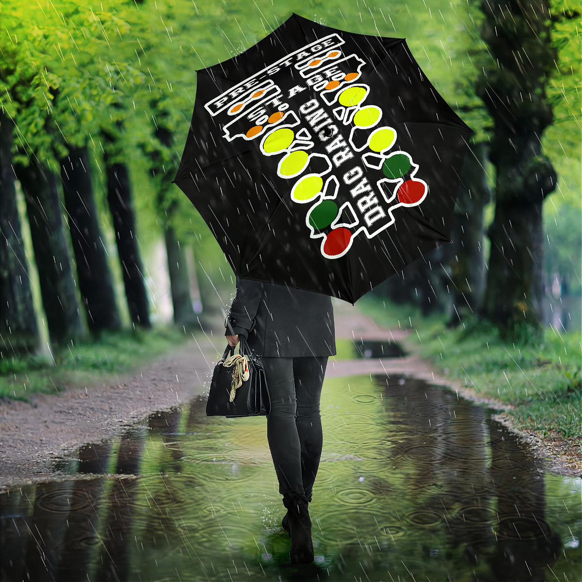 A person walking in the rain along a tree-lined path, holding a black umbrella with a colorful graphic design featuring the words 'TODAY IS A GOOD DAY' and fruit illustrations. The person is wearing a dark coat and carrying a black handbag.