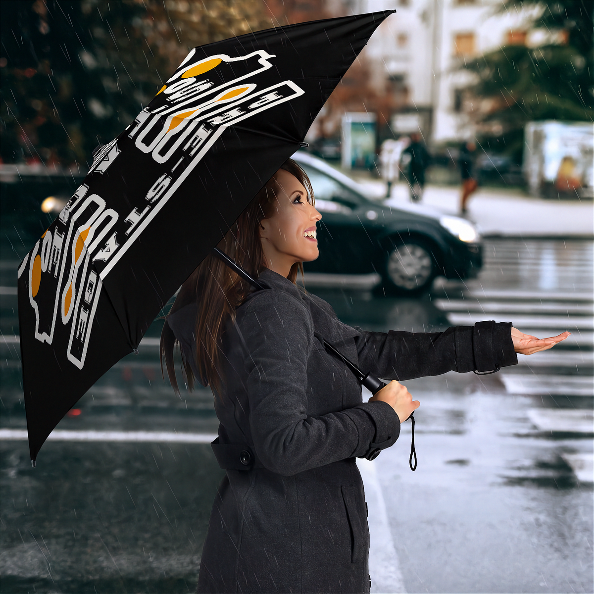 A woman in a dark gray coat stands in the rain holding a black umbrella with a white and yellow graphic design. The background shows a blurred city street scene with wet pavement and a passing car.