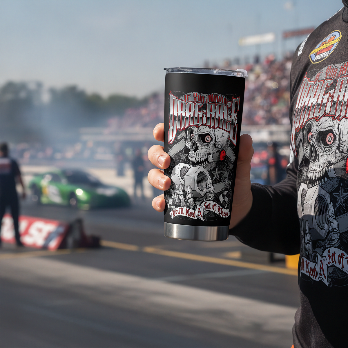 A person at a racetrack holds a black insulated tumbler with a skull and engine graphic that reads 'DRAG RACE'. The person is wearing a matching black long-sleeve shirt with a large skull graphic.