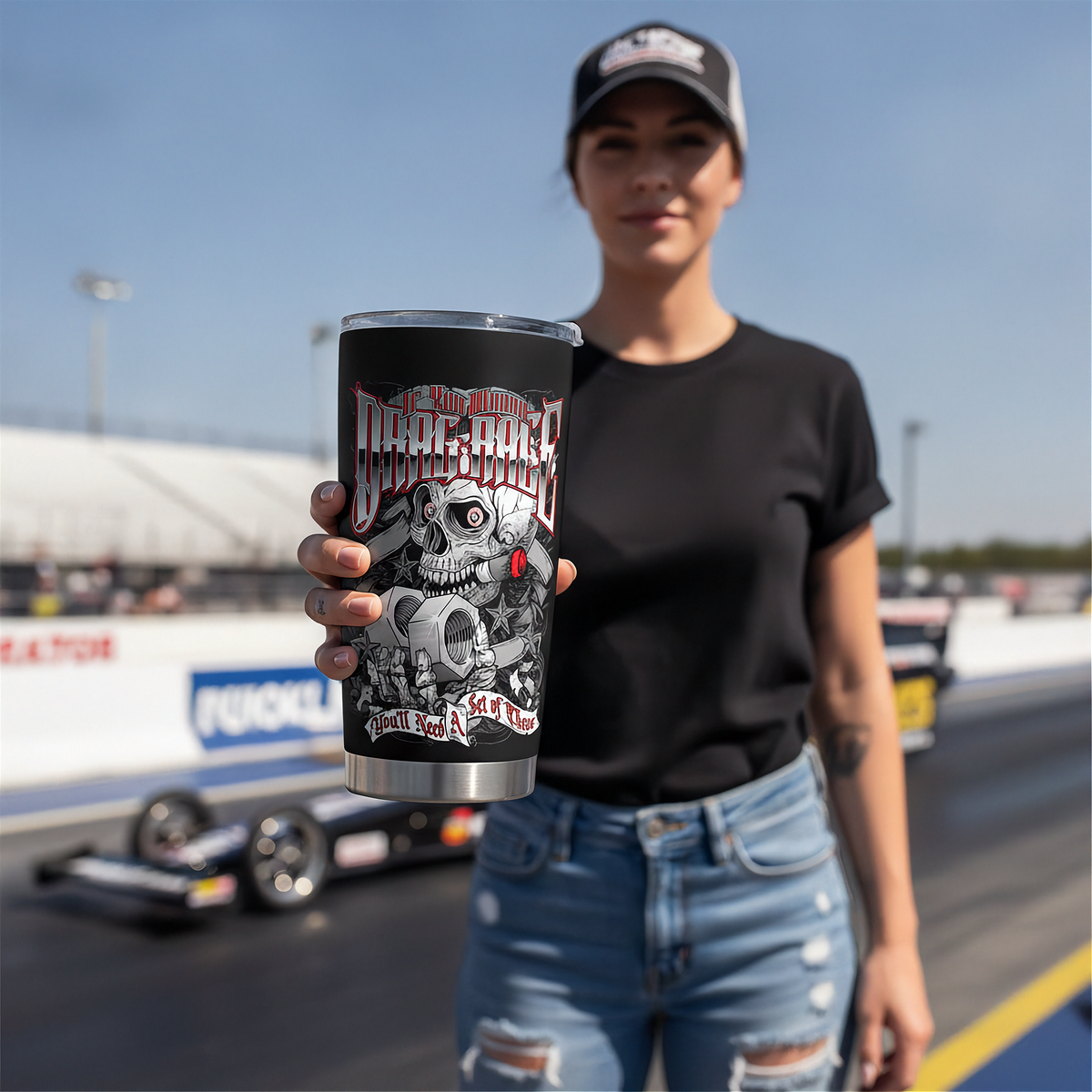 A woman on a race track holds up a black insulated tumbler with a detailed skull and graphic design that reads 'The Drag Race'. The background shows a blurred race car and track barriers under a clear blue sky.