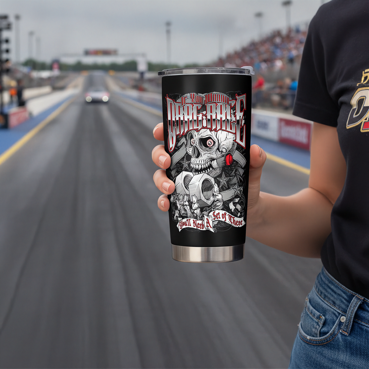 A person holds a black insulated tumbler with a racing graphic in front of a blurred drag strip background. The tumbler features a skull, gears, and the text 'Drag Race'.