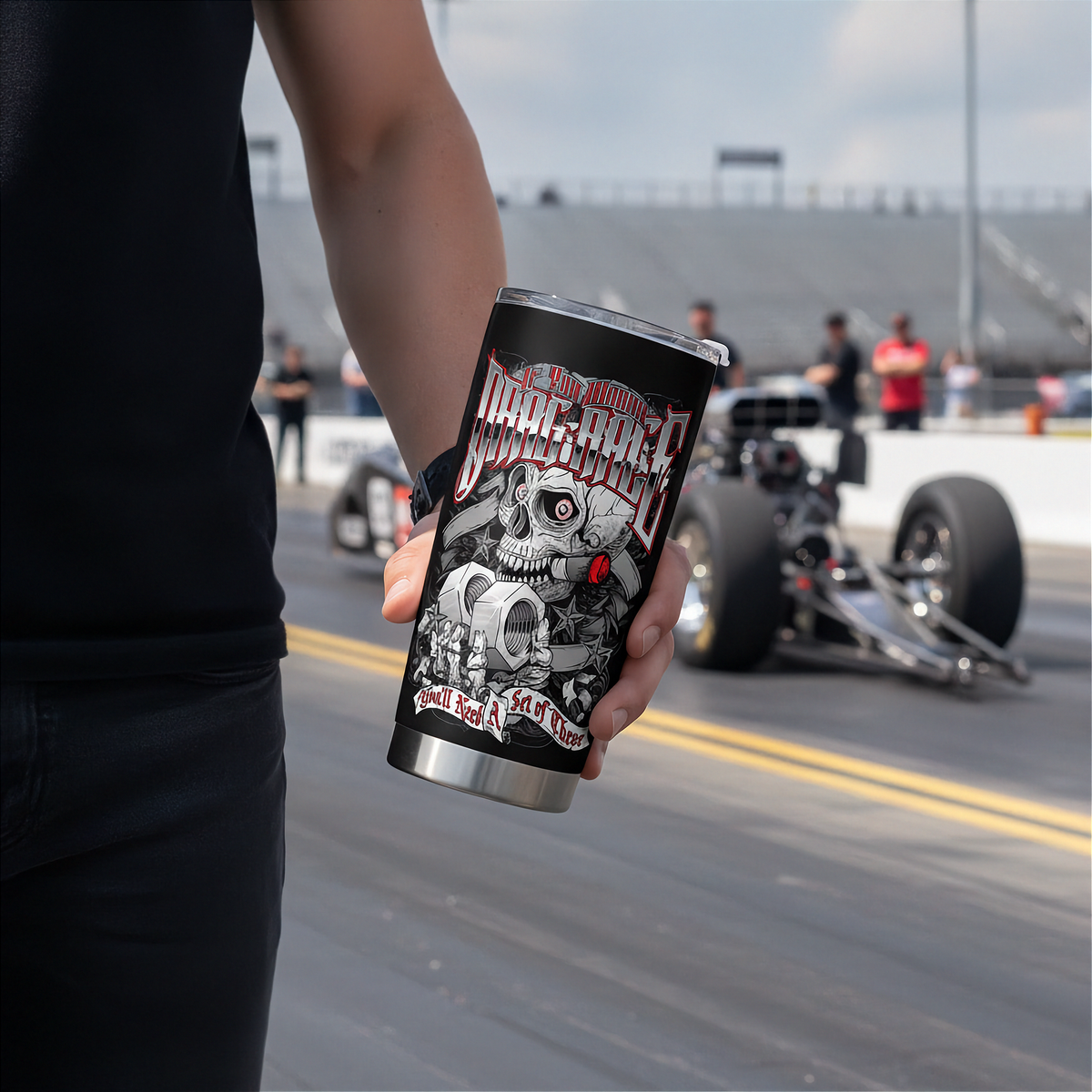 A person in a black shirt holds a black insulated tumbler with a detailed skull and mechanical graphic and the text 'Drag Race' against a blurred racetrack background with a race car.