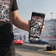 A person at a racetrack holds a stainless steel travel tumbler with a black wrap featuring a skull, engine parts, and the text 'Drag Race'.