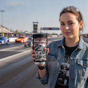 A woman at a racetrack holds up a black insulated tumbler with a graphic design that reads 'If You Wanna To Drag Race You'll Need A Set of These'. She is wearing a denim jacket over a black graphic t-shirt.