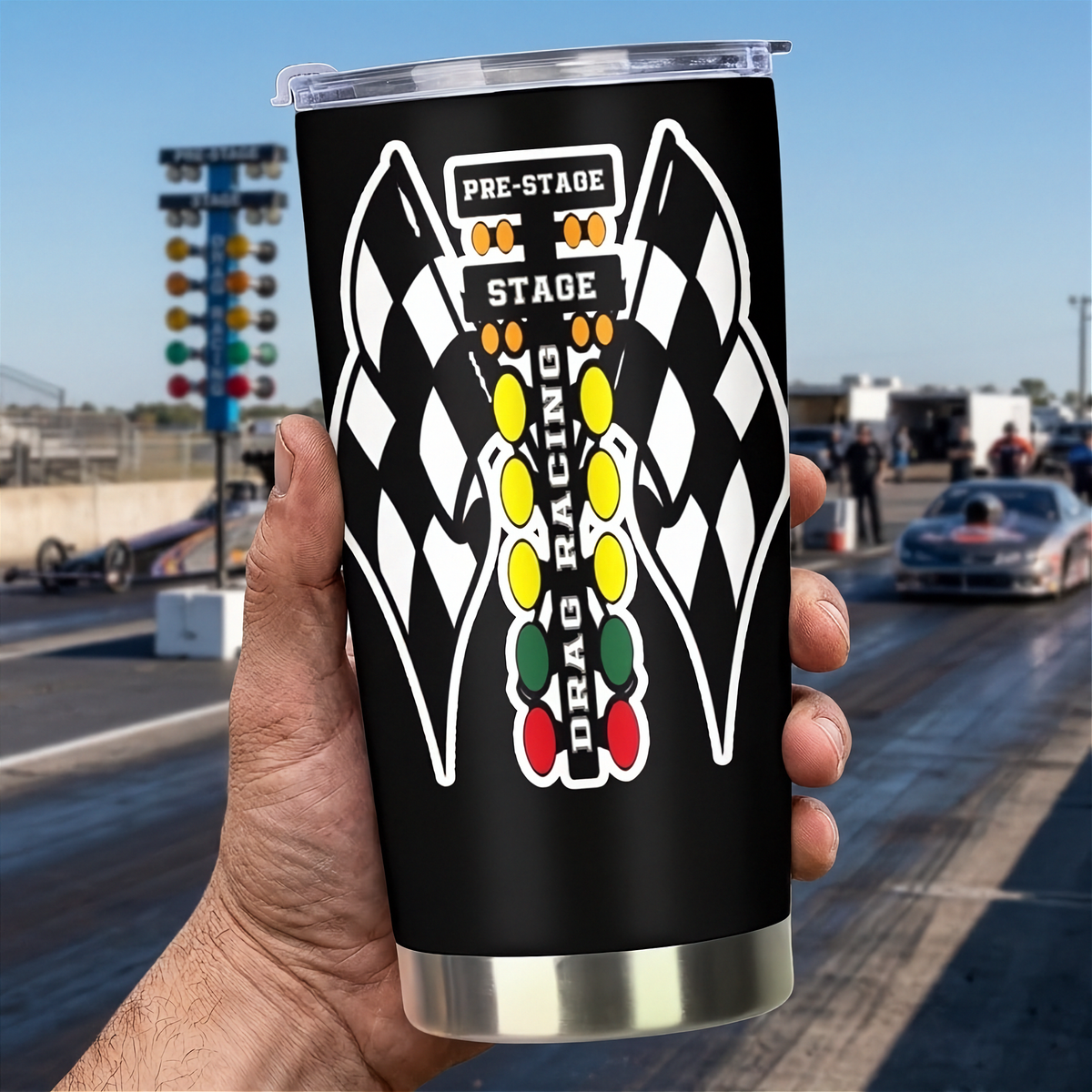 A hand holds a black insulated tumbler with a clear lid and a drag racing graphic featuring checkered flags and a vertical light bar. The background shows a blurred drag strip with a car, track lights, and a blue sky.