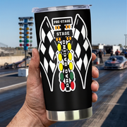 A hand holds a black insulated tumbler with a clear lid and a drag racing graphic featuring checkered flags and a vertical light bar. The background shows a blurred drag strip with a car, track lights, and a blue sky.