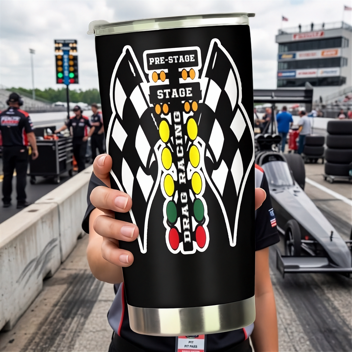 A person at a drag racetrack holds up a black insulated tumbler with a clear lid. The tumbler features a large graphic of two checkered flags facing each other with a vertical column of colored oval lights and the words 'PRE-STAGE', 'STAGE', and 'DRAG RACING'.