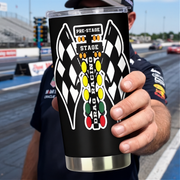 A person at a racetrack holds up a black insulated tumbler with a clear lid. The tumbler features a large graphic of two checkered flags facing each other with a vertical column of colored oval lights and the words 'PRE-STAGE', 'STAGE', and 'DRAG RACING'.
