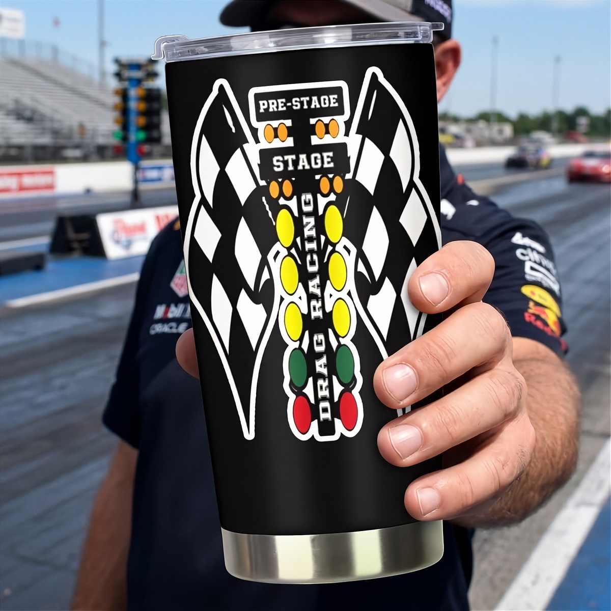 A person at a racetrack holds up a black insulated tumbler with a clear lid. The tumbler features a large graphic of two checkered flags facing each other with a vertical column of colored oval lights and the words 'PRE-STAGE', 'STAGE', and 'DRAG RACING'.