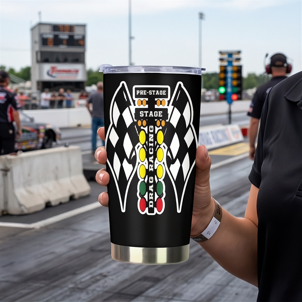 A woman at a drag racetrack holds up a black insulated tumbler with a clear lid. The tumbler features a large graphic of two checkered flags facing each other with a vertical column of colored oval lights and the words 'PRE-STAGE', 'STAGE', and 'DRAG RACING'.