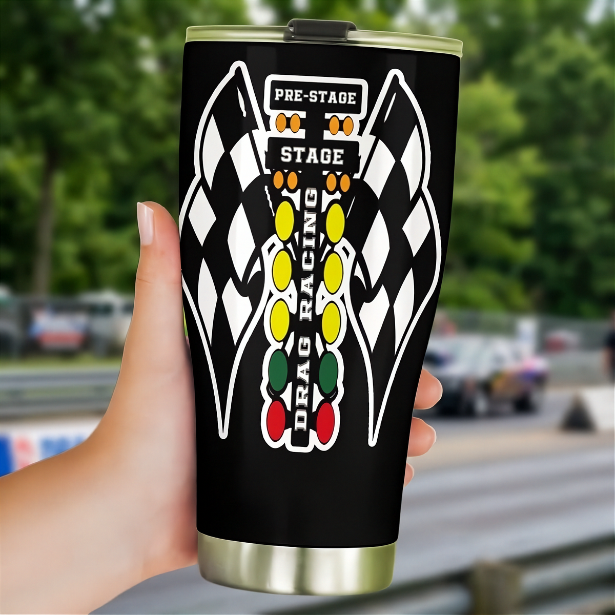 A hand holding a black travel tumbler with a racing-themed graphic that includes checkered flags, the words 'PRE-STAGE', 'STAGE', and 'DRAG RACING', against a blurred racetrack background.