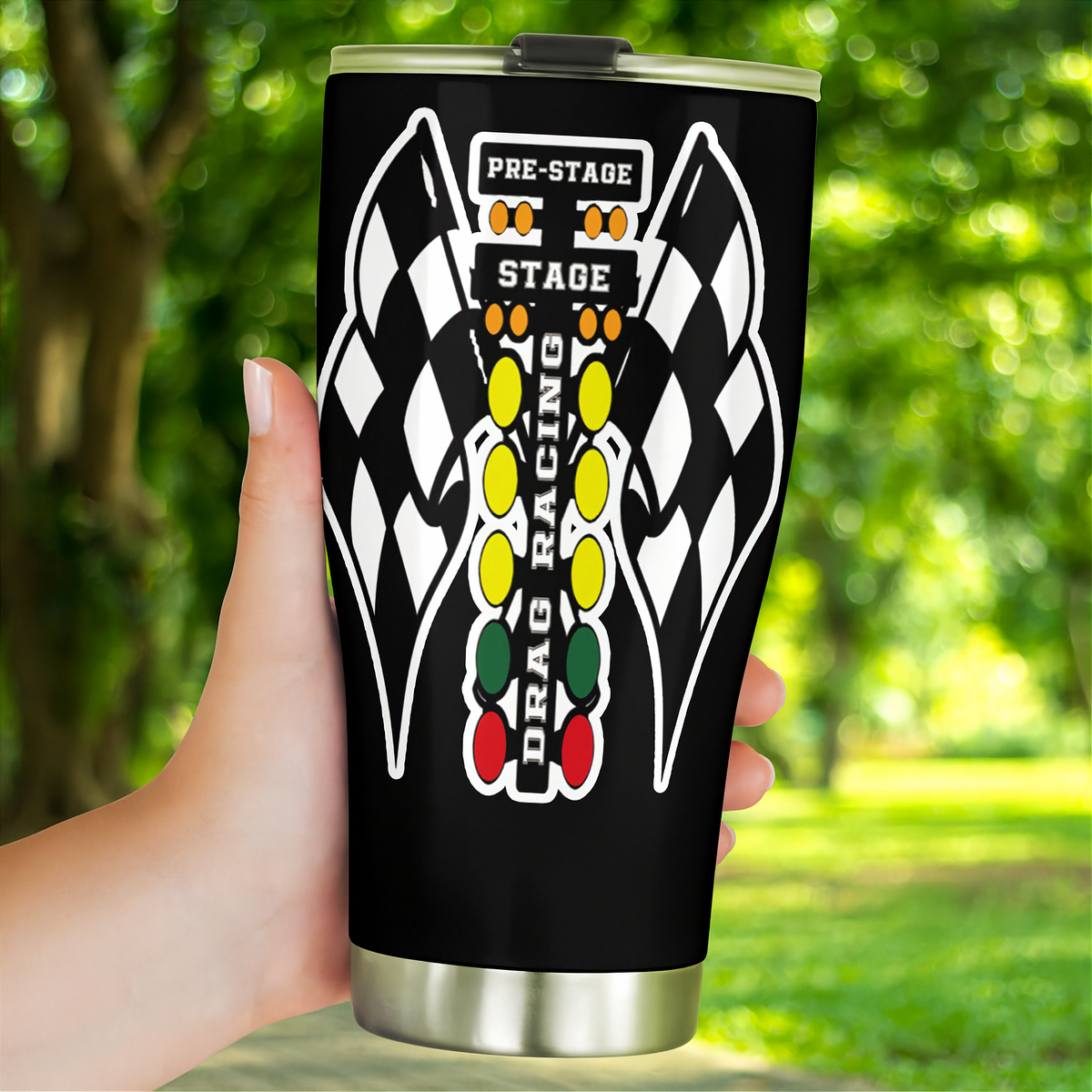 A hand holding a black insulated travel tumbler with a stainless steel lid and base. The tumbler features a white and black checkered wing graphic and a vertical 'DRAG RACING' graphic with traffic light colors (yellow, green, red) on a blurred green outdoor background.