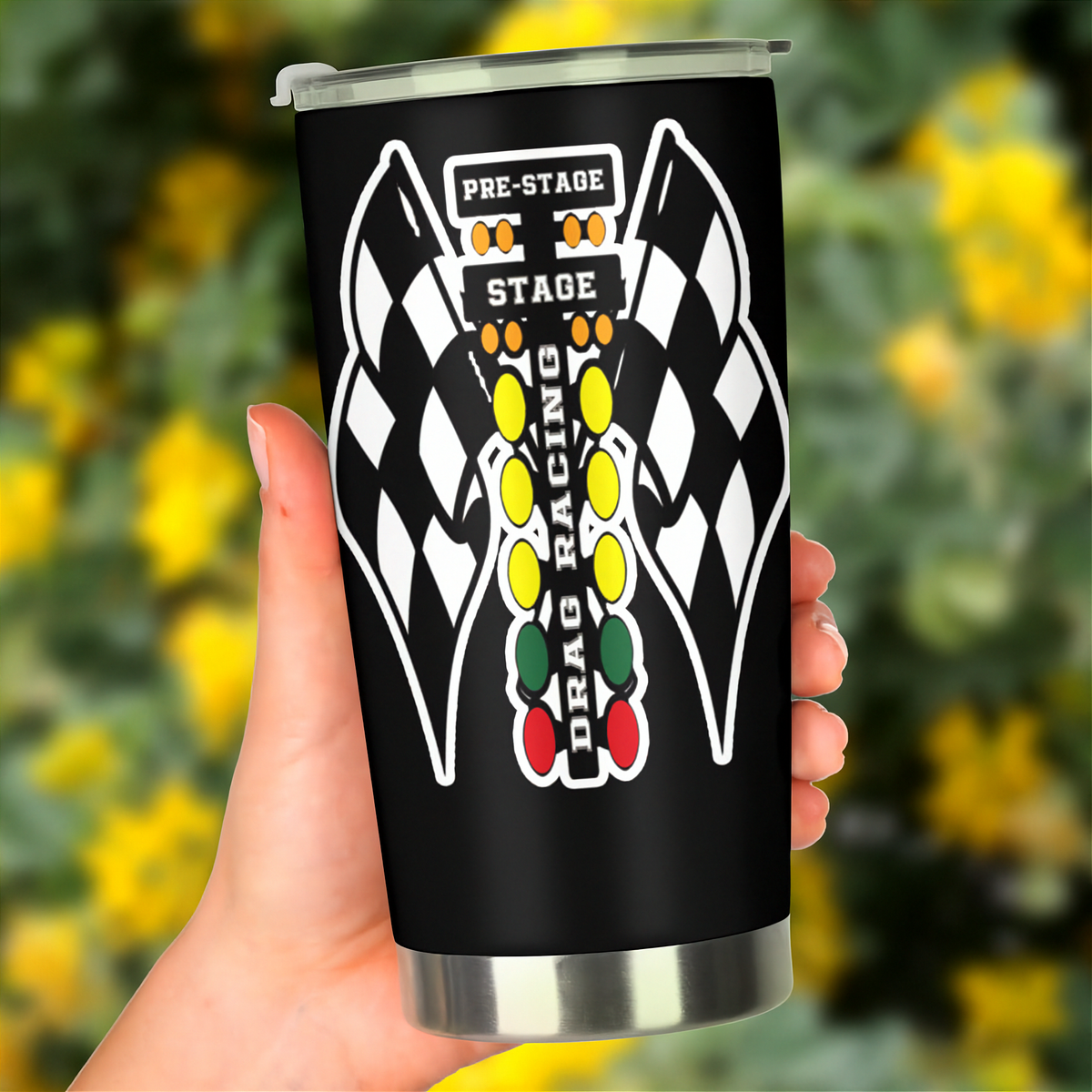 A hand holding a black insulated travel tumbler with a clear lid. The tumbler features a white, yellow, green, and red drag racing graphic with checkered flags and the words 'PRE-STAGE', 'STAGE', and 'DRAG RACING'.