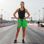 A woman stands on a racetrack wearing bright green athletic shorts with a black drawstring and a white graphic logo on the left leg, paired with a black tank top and black sneakers.