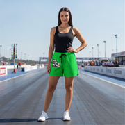 A woman stands on a drag strip wearing bright green athletic shorts with a black drawstring and a graphic print on the left leg, paired with a black tank top and white sneakers.