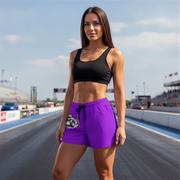 A woman stands on a racetrack wearing a black sports bra and bright purple athletic shorts with a white and black graphic print on the left leg.