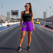 A woman stands on a drag racing track wearing a black tank top and bright purple athletic shorts with a graphic print on the left leg. The background shows the racetrack, track lights, and grandstands under a clear sky.