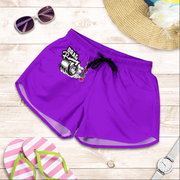 Purple athletic shorts with a black drawstring and a 'DRAG Racing' graphic on the left leg, displayed in a flat-lay arrangement on a light wood background with sunglasses, a straw hat, flip-flops, and a watch.