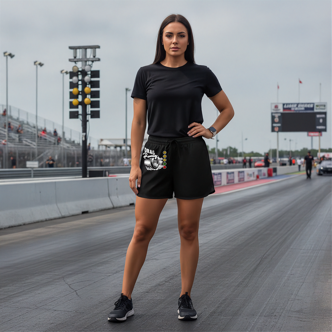 A woman stands on a drag racing track wearing a black short-sleeve t-shirt and black athletic shorts with a white and black graphic print on the left leg, paired with black sneakers.