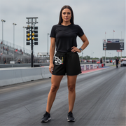 A woman stands on a drag racing track wearing a black short-sleeve t-shirt and black athletic shorts with a white and black graphic print on the left leg, paired with black sneakers.
