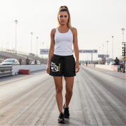 A woman walking on a racetrack wearing black athletic shorts with a white graphic print, a white tank top, and black sneakers.
