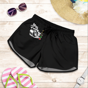 Black women's shorts with a white and green 'Trash Panda' graphic, styled in a flat lay on a light wood background with sunglasses, a straw hat, pink flip-flops, and a watch.