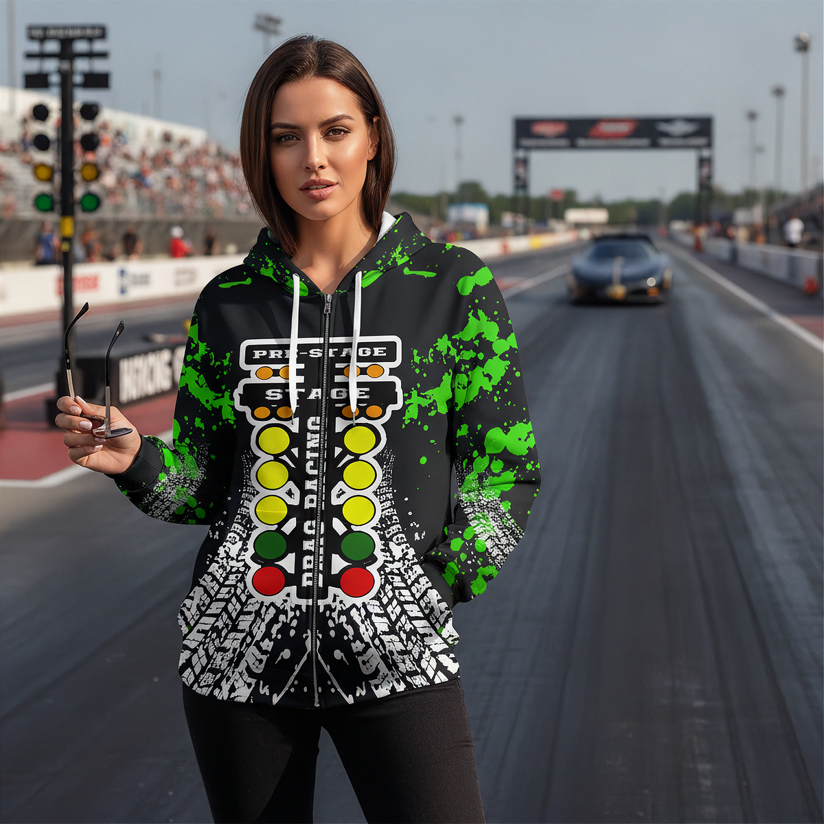 A woman stands on a racetrack wearing a black zip-up hoodie with a green paint-splatter design and a large racing light graphic on the front. A car is visible in the background of the racetrack.