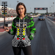 A woman stands on a racetrack wearing a black zip-up hoodie with a green paint-splatter design and a large racing light graphic on the front. A car is visible in the background of the racetrack.