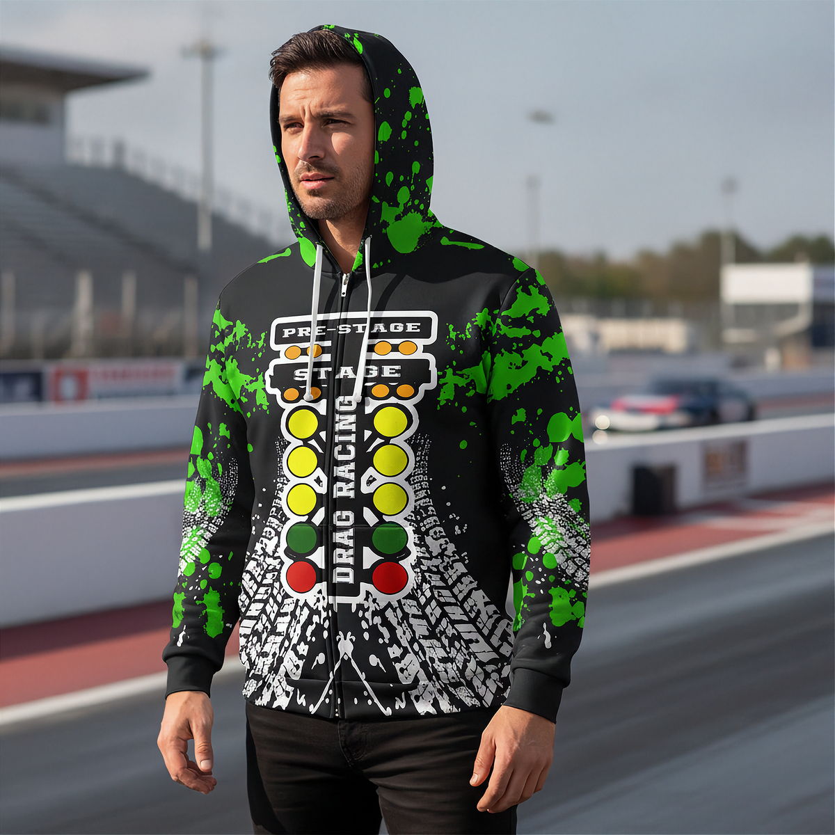 A man wearing a black zip-up hoodie with a green paint splatter design and a drag racing theme graphic on the front, standing on a racetrack.