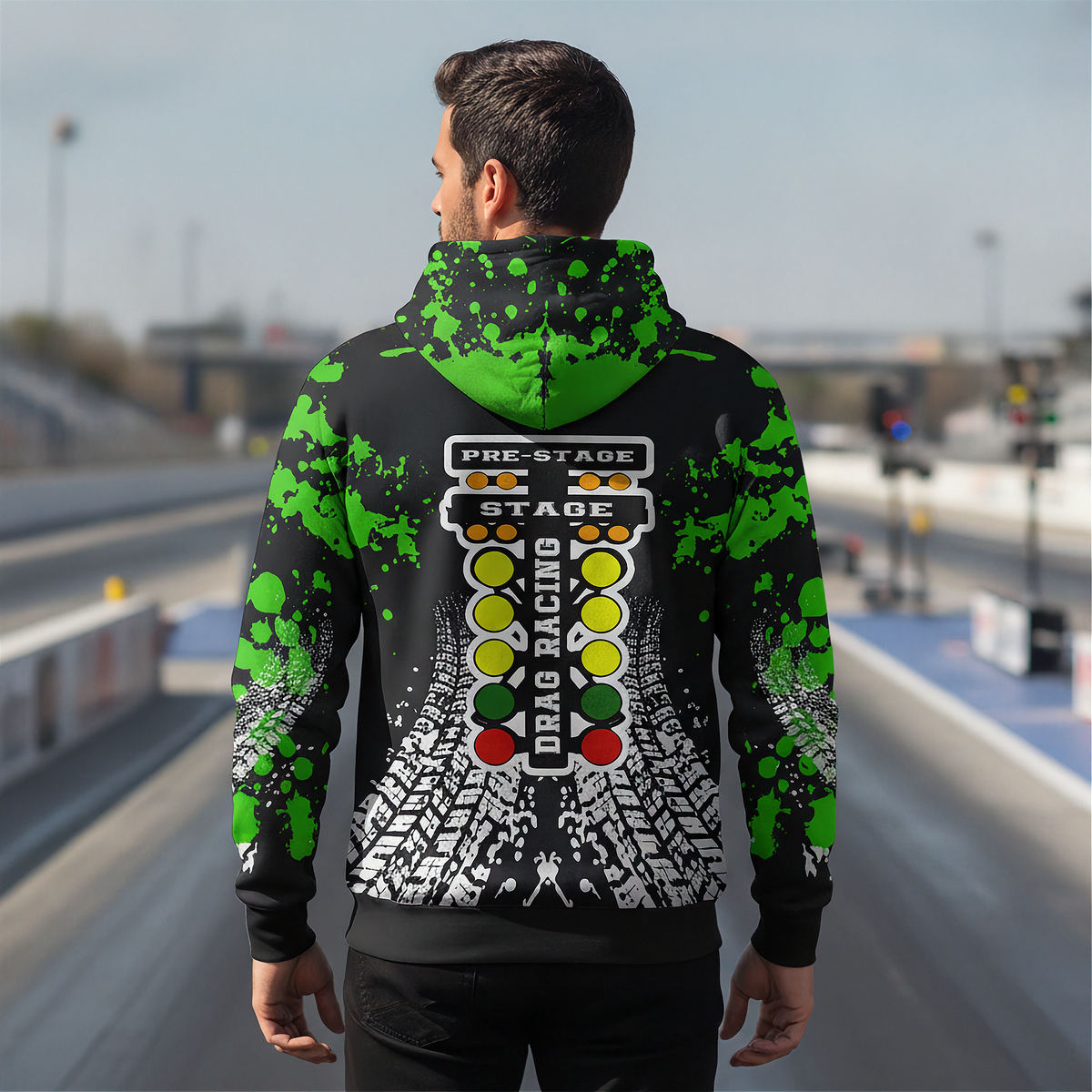 A man viewed from behind wearing a black hoodie with a green splatter pattern and a large racing-themed graphic on the back, standing on a racetrack.