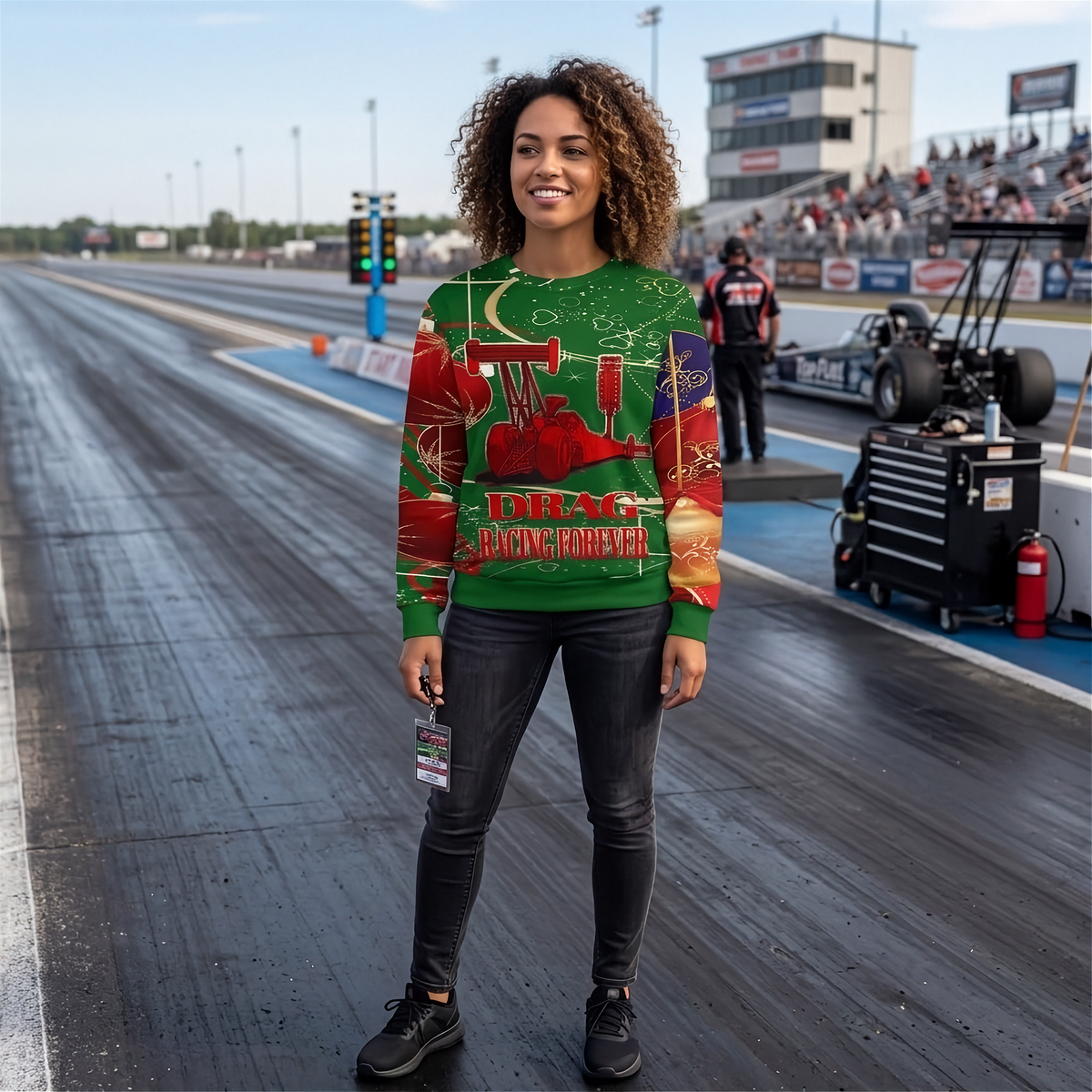 A woman stands on a drag strip wearing a green crewneck sweatshirt with a red and gold 'Racing for Victory' graphic, paired with black skinny jeans and black sneakers.