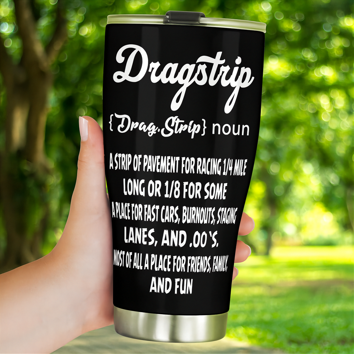 Hand holding a black Dragstrip Noun Tumbler outdoors against a leafy green background.