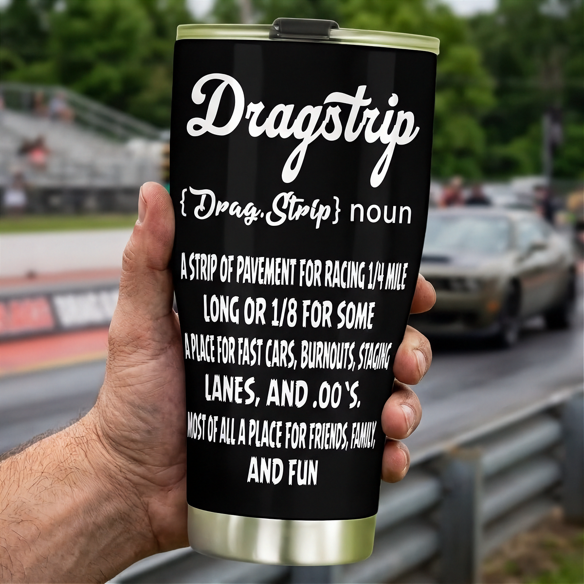 Hand holding a black Dragstrip Noun Tumbler with white definition lettering beside a drag racing track.