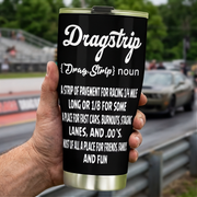 Hand holding a black Dragstrip Noun Tumbler with white definition lettering beside a drag racing track.