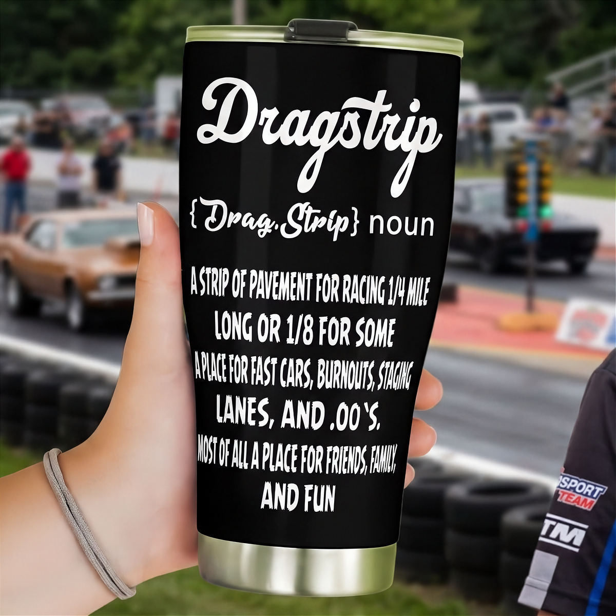 Black Dragstrip definition tumbler held near the starting line, with race cars and drag tree lights behind it.