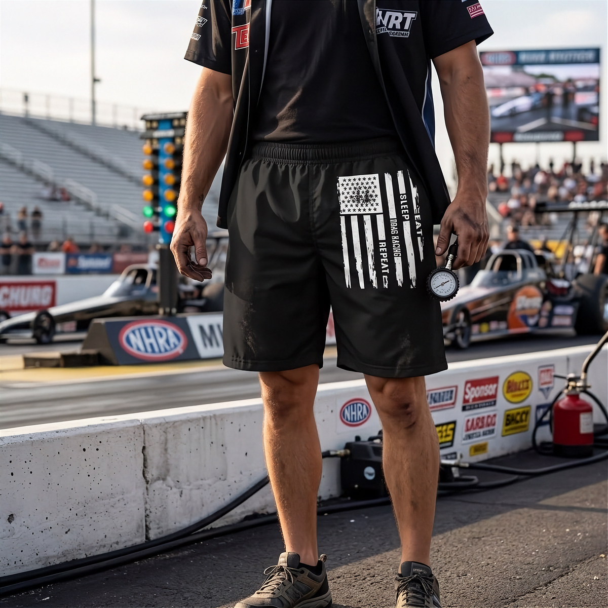A person standing at a drag racing track wearing black shorts with a vertical American flag graphic and white text reading 'EAT SLEEP DRAG RACING REPEAT' on the left leg, along with a black shirt and vest in a pit area with race cars and track signage in the background.