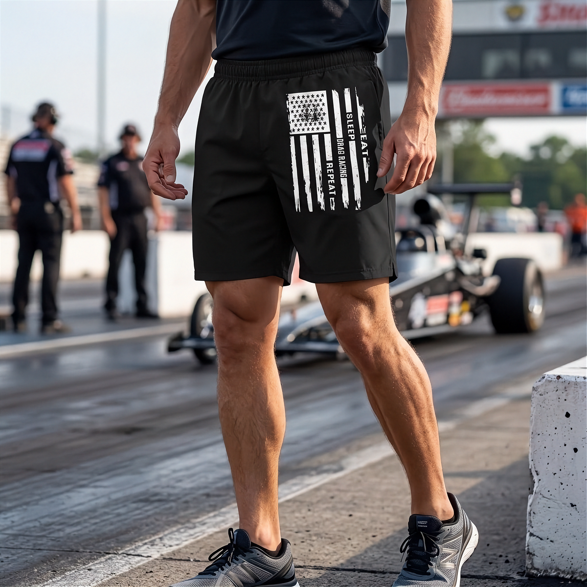 A man standing on a drag racing track wearing black athletic shorts with a distressed white American flag graphic on the left leg. A drag race car is blurred in the background on the track.