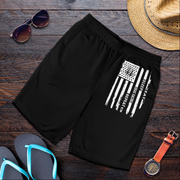 Black athletic shorts with a distressed white American flag graphic on the left leg, styled in a flat lay on a dark wood background with sunglasses, a straw hat, flip-flops, and a wristwatch.