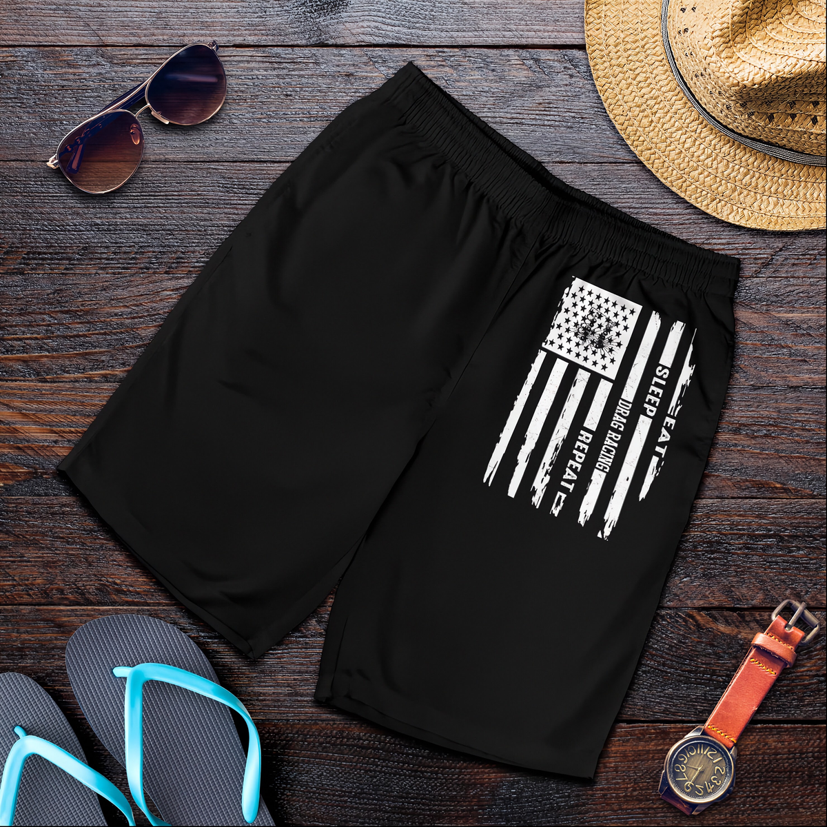 Black athletic shorts with a distressed white American flag graphic on the left leg, styled in a flat lay on a dark wood background with sunglasses, a straw hat, flip-flops, and a wristwatch.