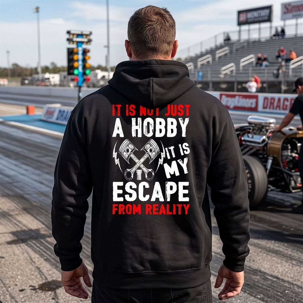 A man viewed from behind wearing a black hoodie with a large back graphic that reads 'IT IS NOT JUST A HOBBY IT IS MY ESCAPE FROM REALITY' in white and red text with crossed pistons. The background shows a drag racing track with grandstands and a race car.