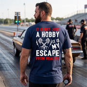 A man standing on a drag strip wearing a navy blue t-shirt with a graphic print on the back. The print features crossed pistons and the text 'IT IS NOT JUST A HOBBY IT IS MY ESCAPE FROM REALITY' in white and red.